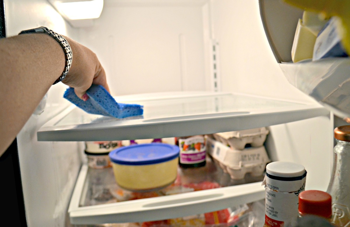 hand holding a blue sponge and wiping down a refrigerator shelf with vinegar