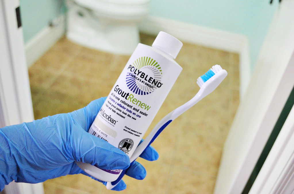 hand wearing blue glove holding white bottle of grout renew and toothbrush 