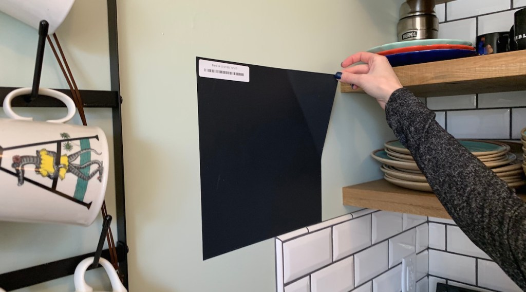 hand holding a black square wall sticker in kitchen with tile backsplash and mugs