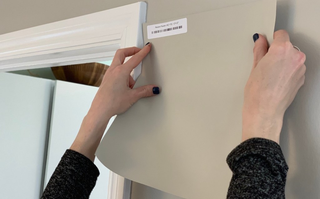 two hands holding a revere pewter paint sample on gray wall with white trim