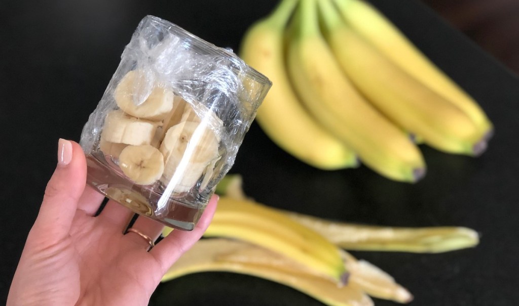 hand holding sliced banana in a glass covered with plastic wrap with bananas in the background