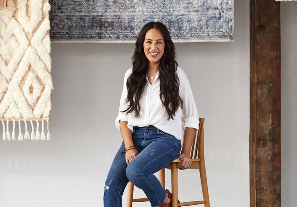 joanna gaines sitting on stool with hanging rugs in the background