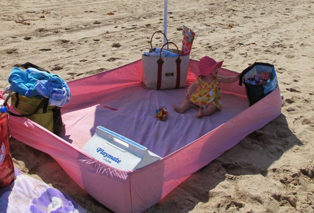 pink fitted sheet in sand with cooler beach bags and baby sitting inside