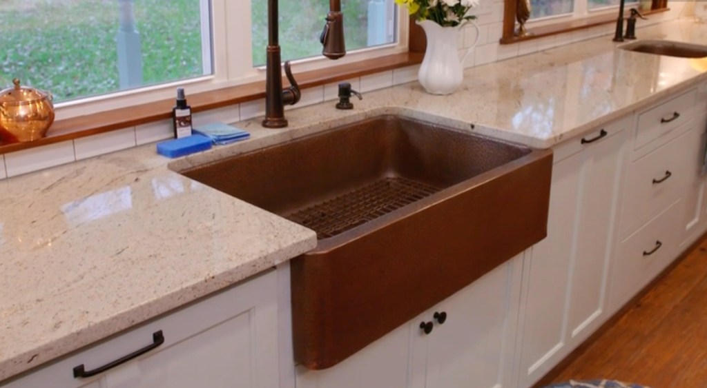 copper apron sink with white cabinets and granite stone countertop below window 