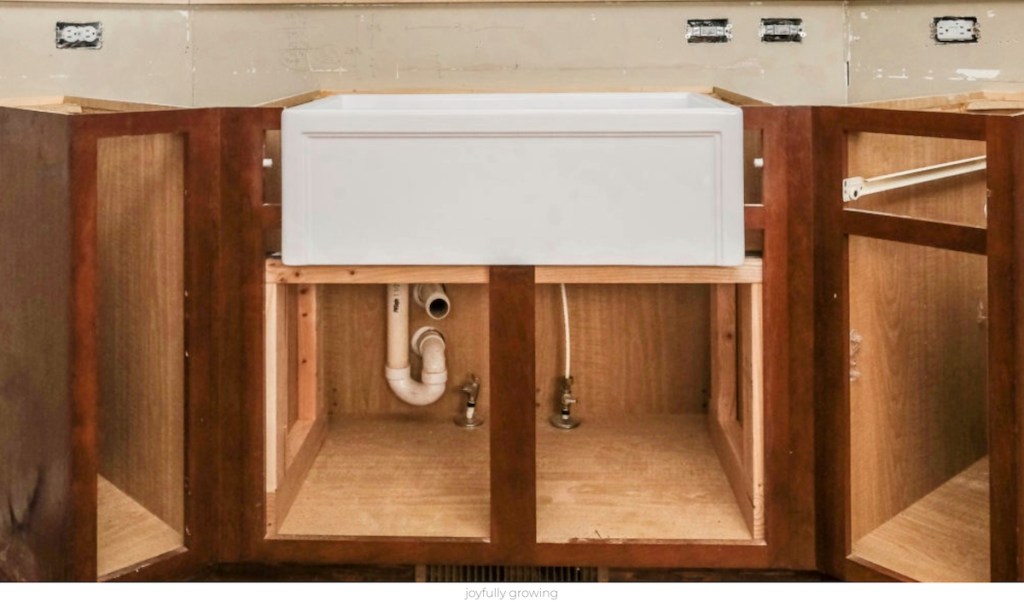 brown kitchen cabinets under construction with doors off and white farmhouse sink sitting in the middle