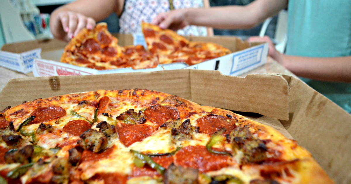 a close up of a 3 topping dominos pizza with kids grabbing slices of a pepperoni pizza in the background