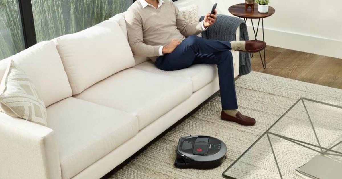 man sitting on a couch while a robotic vacuum cleans near his feet