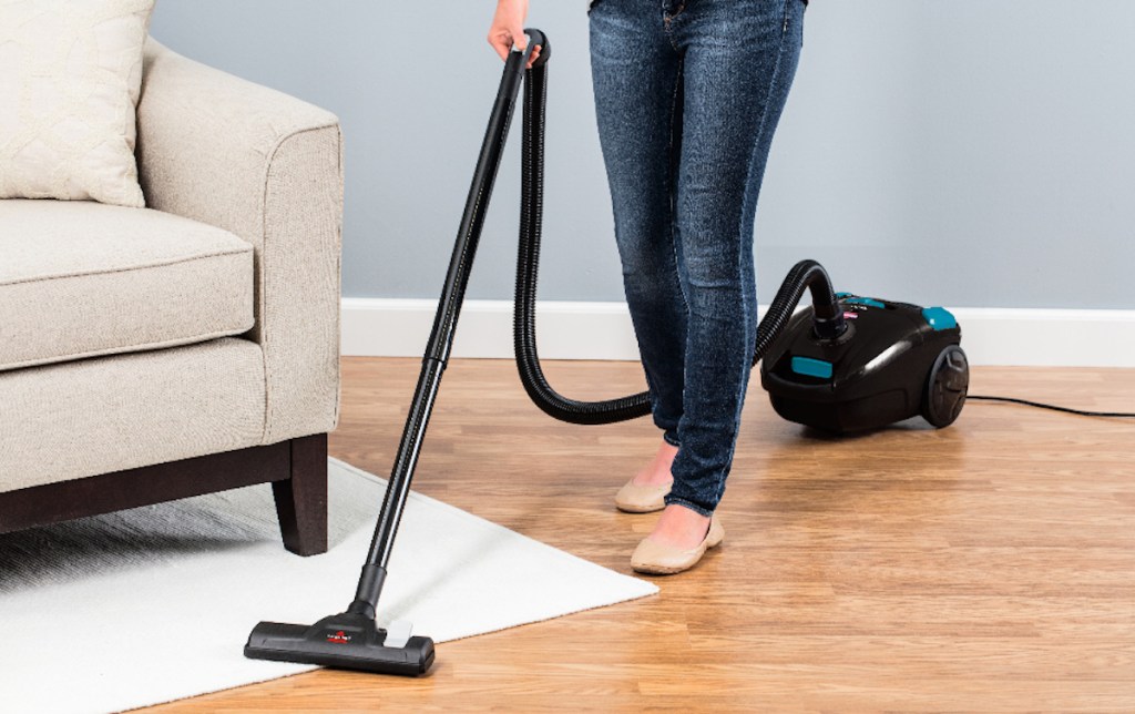 person holding arm of black vacuum next to cream beige couch and rug in living room