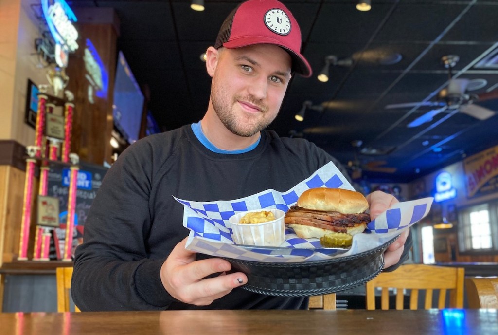 man sitting at table smiling holding a sandwich in basket