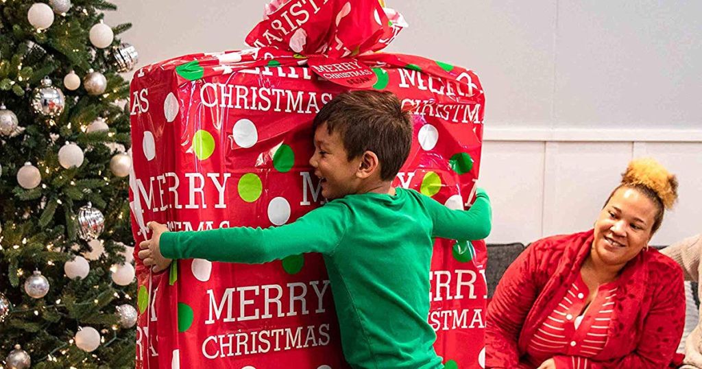 little boy hugging large wrapped gift with christmas tree and adult