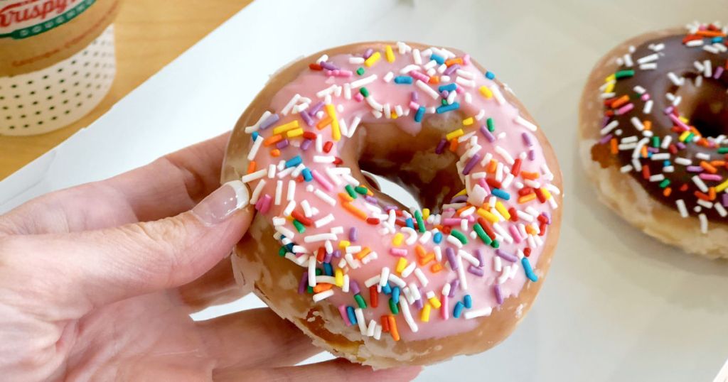 Womans hand with french manicure holding strawberry sprinkle krispy kreme donut with chocolate sprinkle donut and coffee in the background