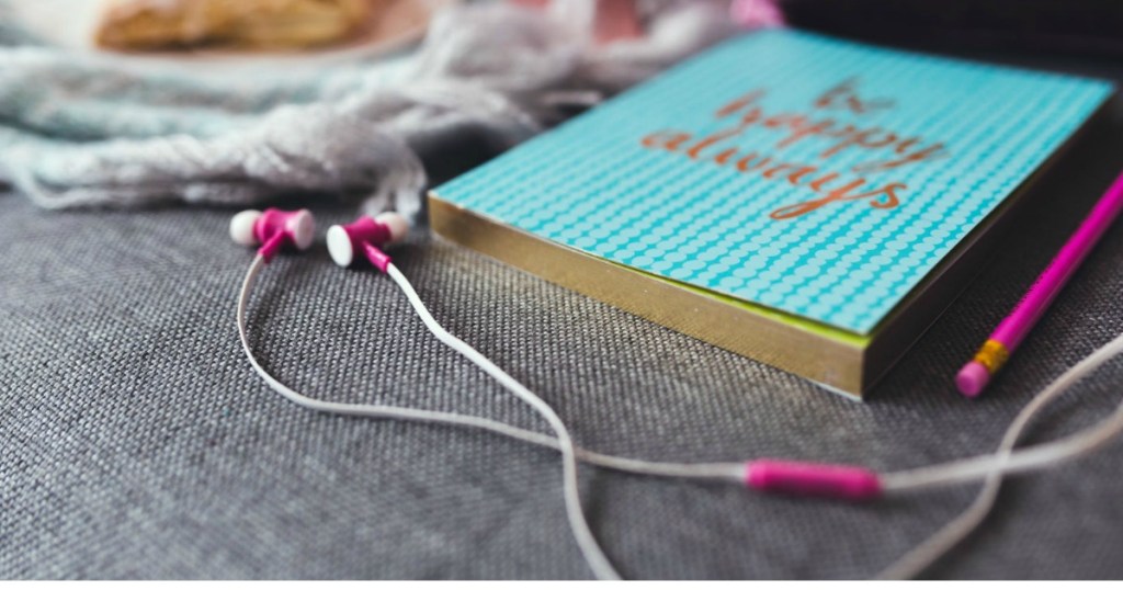 pink and white earbuds laying on cushion next Be Happy Always planner