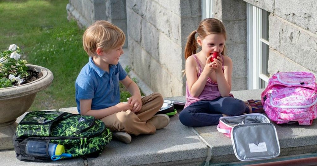 boy and girl sitting on ledge at school with backpacks girl is eating apple