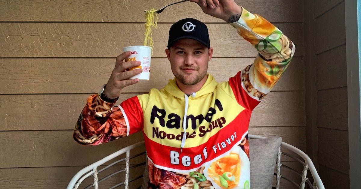 man wearing a ramen noodles hoodie holding a cup of noodles next to his head with the noodles stretching from fork