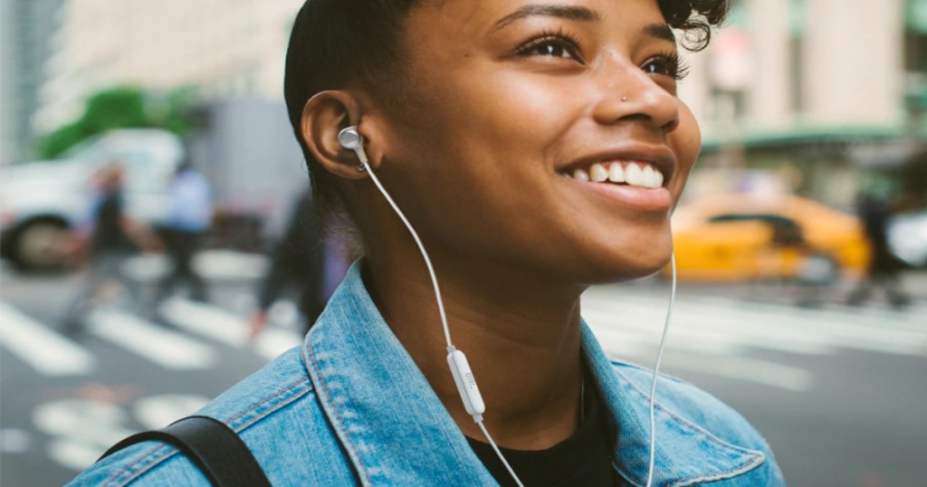 A lady outside walking in town wearing JBL in ear headphones, smiling