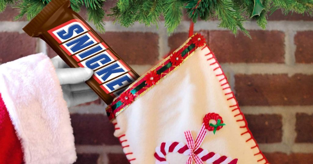 Santa hand putting giant snickers into stocking hanging on brick mantle with garland