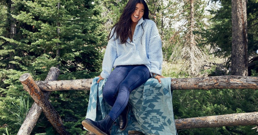 woman in blue leggings and sherpa top sitting on a log fence outside