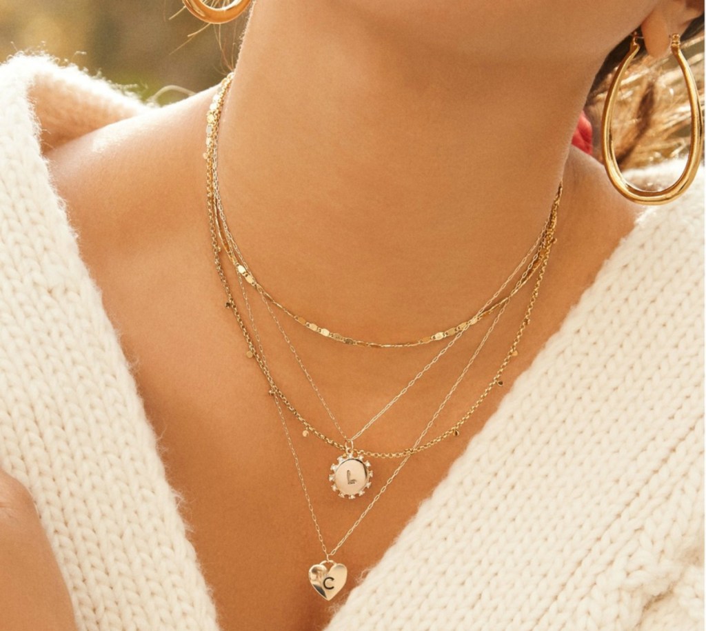 woman in white sweater wearing multiple gold initial pendant necklaces