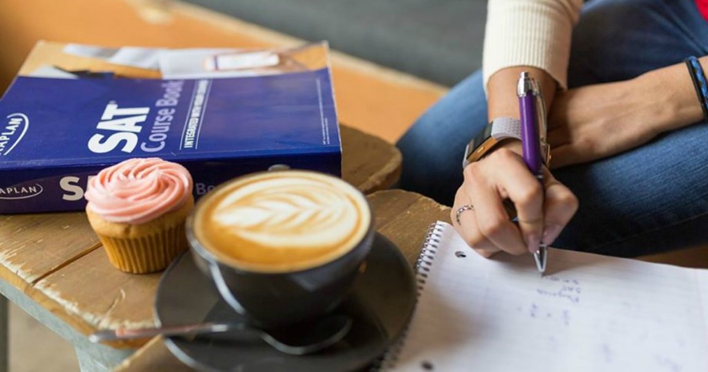 person writing in notebook next to SAT prep book