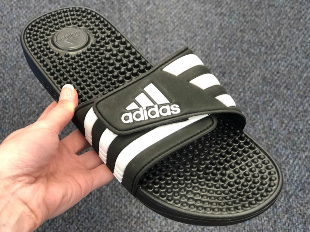 Single of men's slip on sandal in black with white adidas logo in hand near carpeted floor