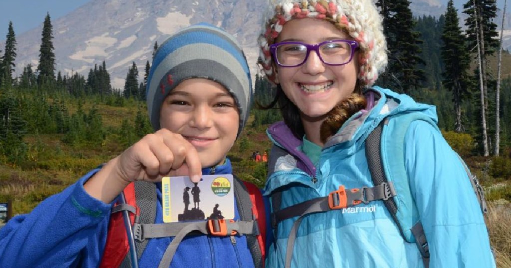 2 children standing next to each other wearng winter gear and one holding a park pass in his hand