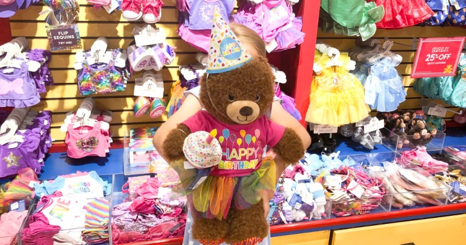 Young girl holding a large Birthday-themed teddy bear in front of her in a store