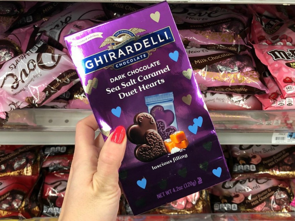 Hand holding a purple bag of gourmet chocolates near in-store display of Valentine's Day candy clearance