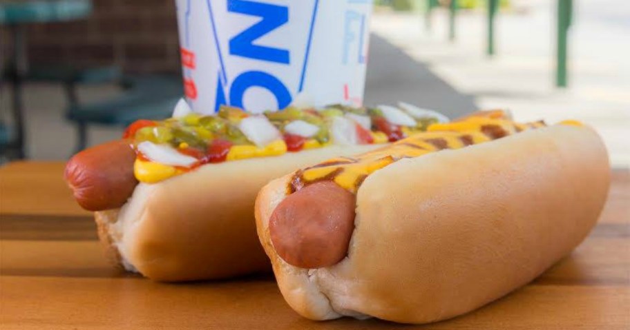Two hot dogs on picnic table near large drink in white cup