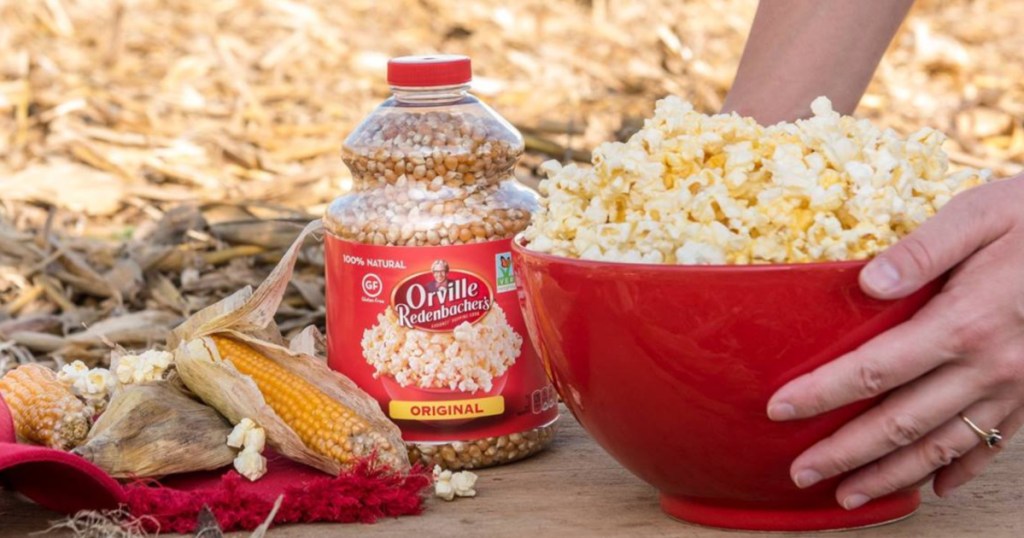 woman holding bowl of popcorn and jar of popcorn kernels and corn on table outside