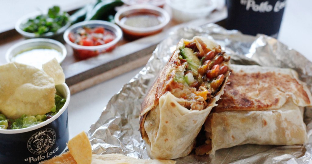 foil laid out with el pollo loco burrito sliced in half on top of it with inside of burrito shown, guacamole with a chip in it on the side, cup of pico de giao in the background with fountain drink