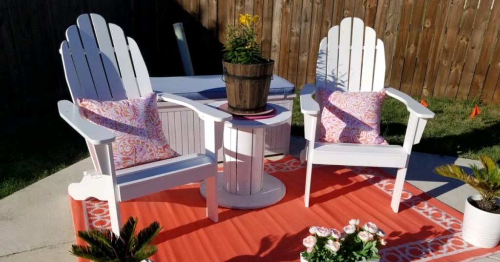 two white Adirondack chairs with pillows, orange rug, and plants outside