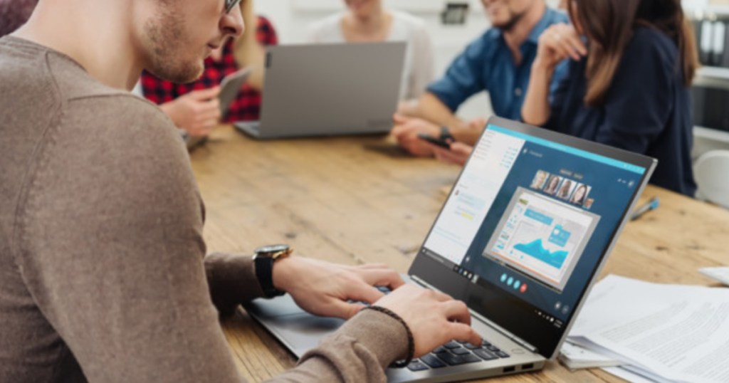 man working on laptop at a brown table with other people in the distance