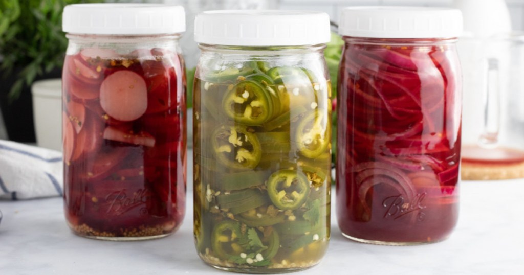 three clear jars with white lids with pickled red and green foods inside