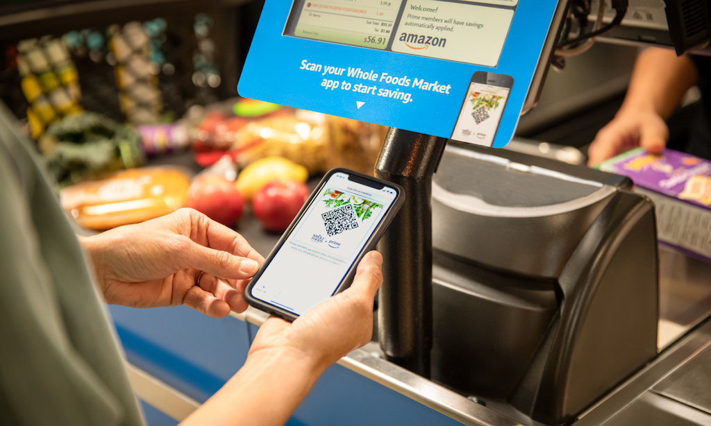 A person holding their phone with the Whole Foods app displayed in front of a Whole Foods register