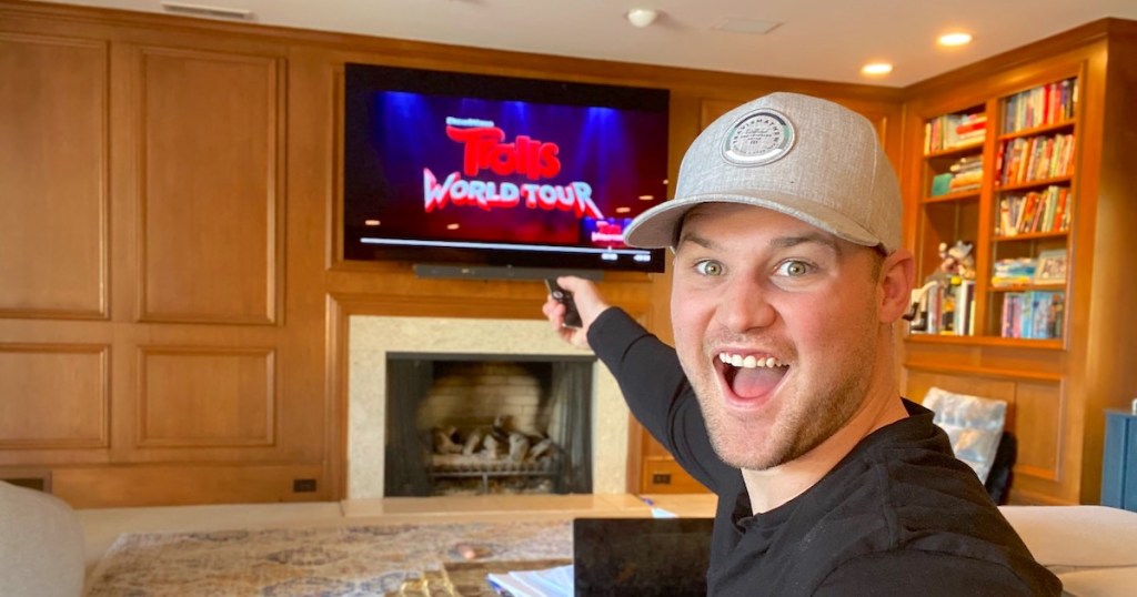 man with surprised happy face holding remote with trolls world tour on screen