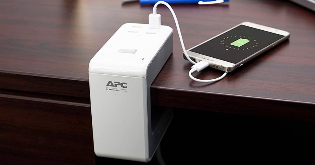 white u-shaped surge protector clipped on to edge of desk with phone plugged in and charing