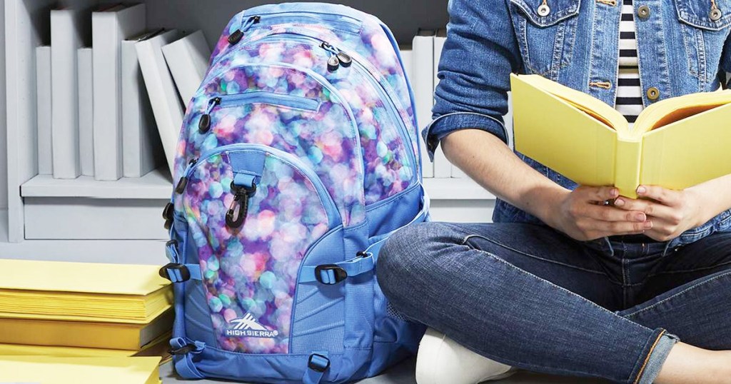 girl sitting on library floor reading a yellow book with blue and pink bubble print backpack next to her