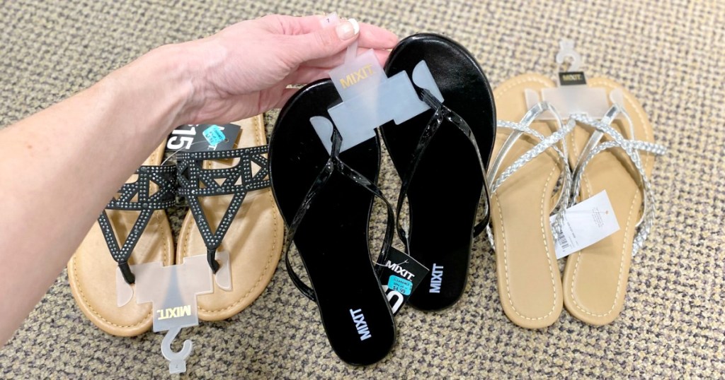 twoman holding up pair of black flip-flops with two other pairs of womens sandals on the floor in the background