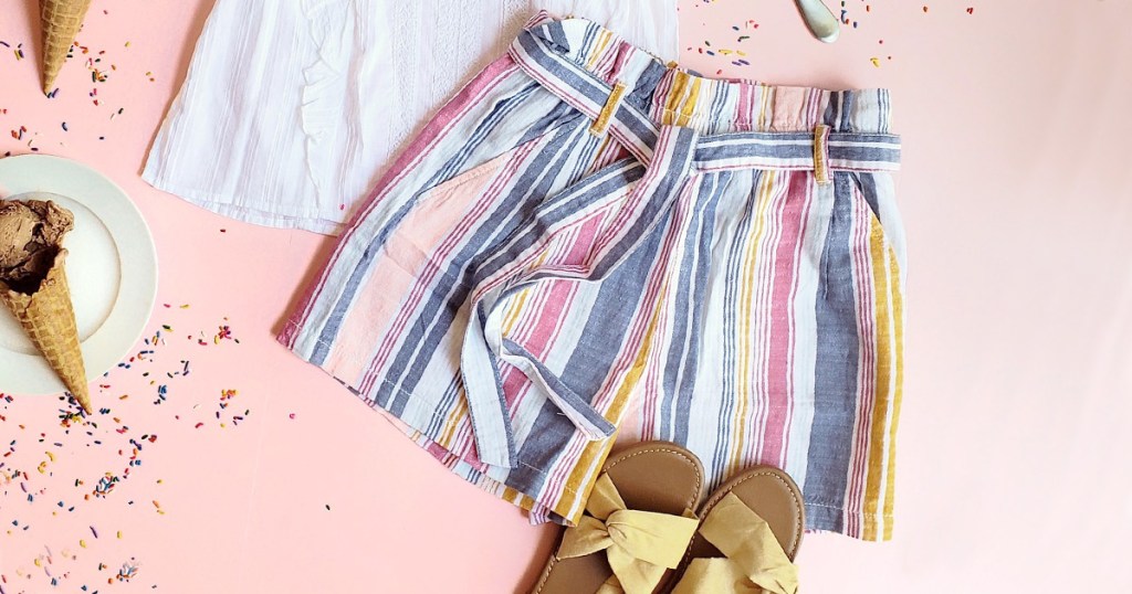 blue, pink, and white striped shorts with tie at waist laying on pink background with white top and yellow sandals