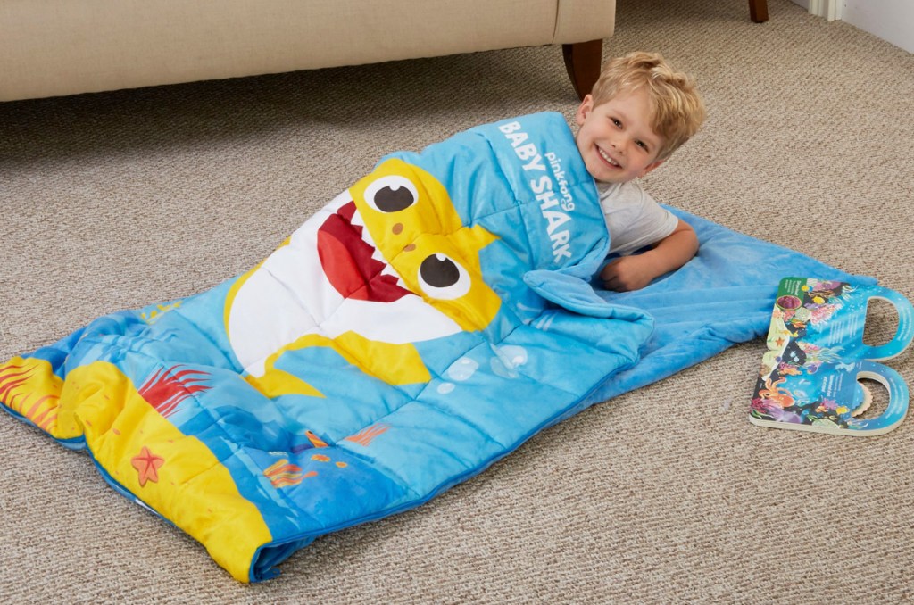 toddler boy laying on floor inside blue baby shark printed sleeping bag with book