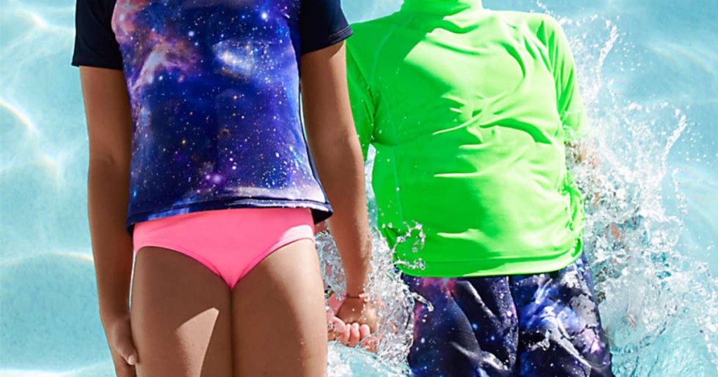 girl in purple and blue galaxy top and pink swim bottoms and boy in blue swim trunks and green rash guard holding hands and jumping into pool