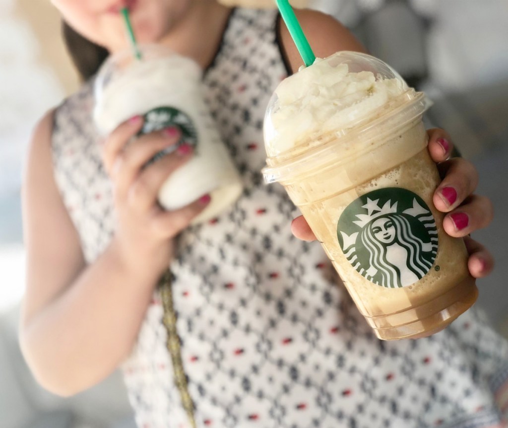 girl with two starbucks frappuccinos, drinking one while holding other out to camera