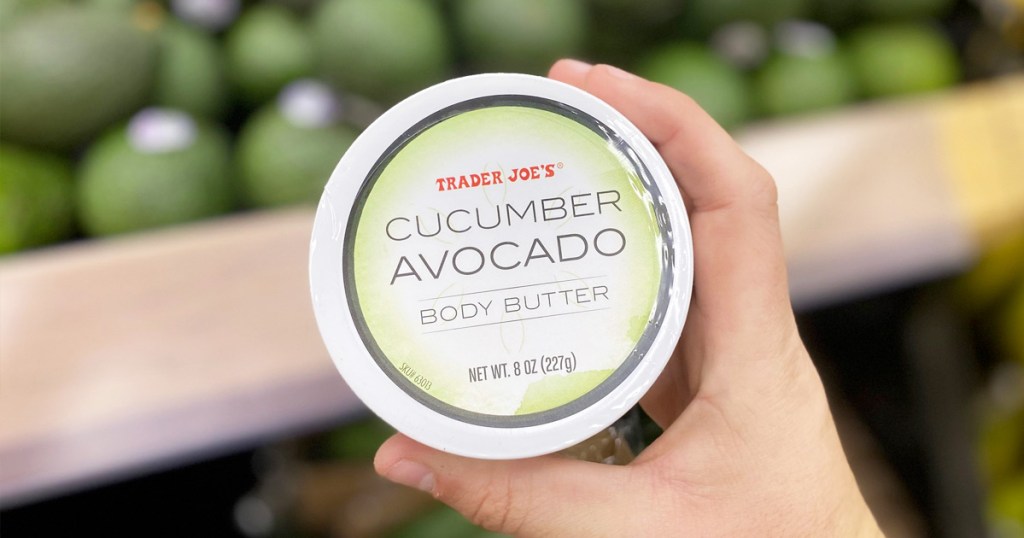 person holding a round container of Trader Joe's Cucumber Avocado Body Butter near avocado display in store