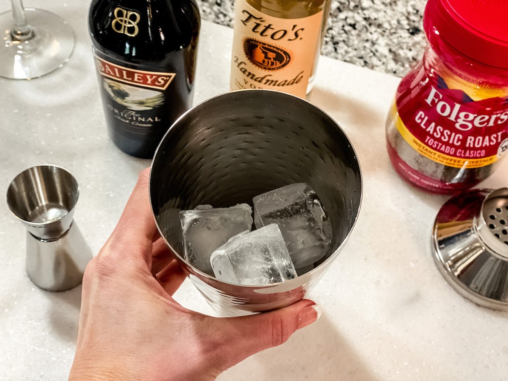 hand holding a stainless steel shaker with ice and ingredients in background