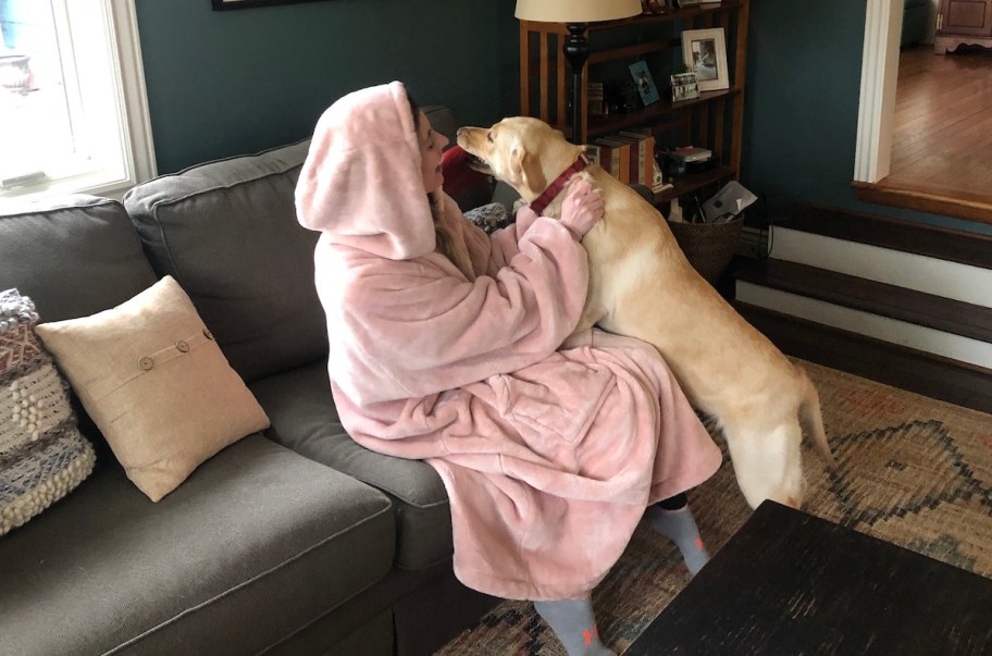 person wearing light pink wearable blanket sitting on couch playing with dog