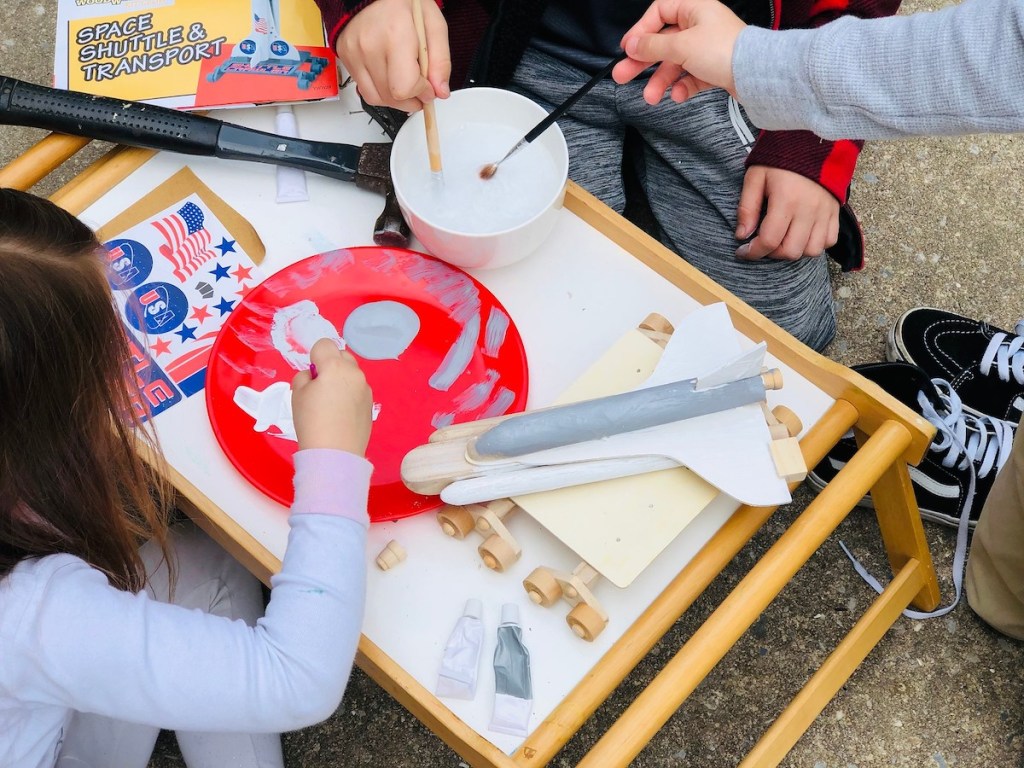 kids with paintbrushes painting wooden rocket ship on tray table