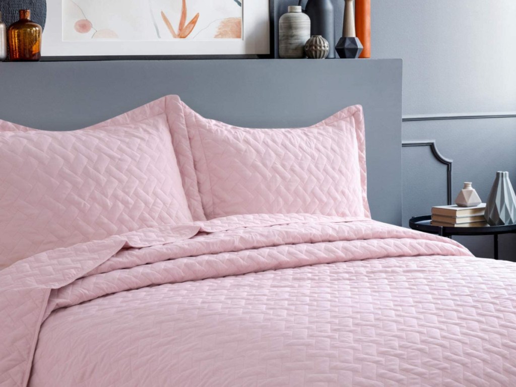 light pink quilt set on a grey bedframe next to a side table with books and nicknacks