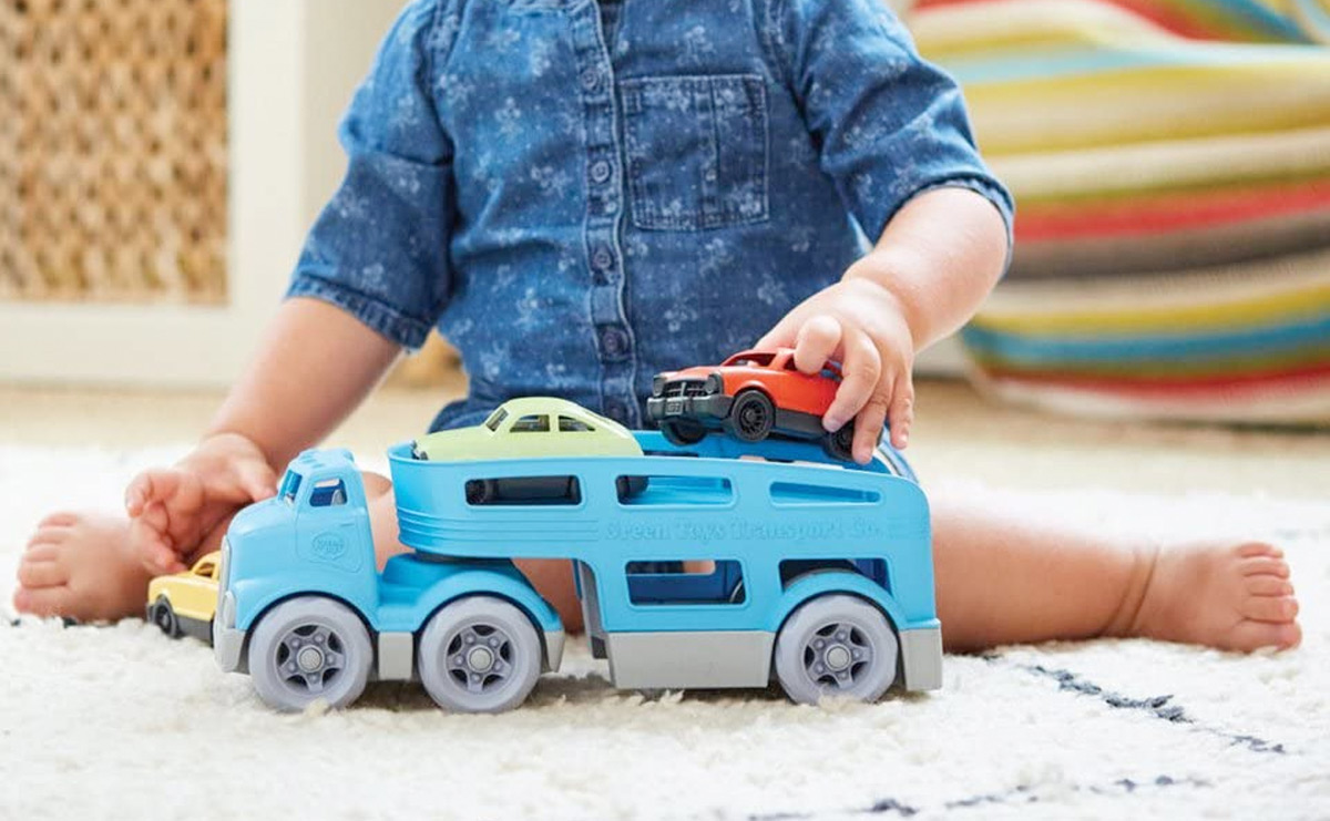 child sitting on rug playing with blue car carrier toy with multiple colors of cars on top