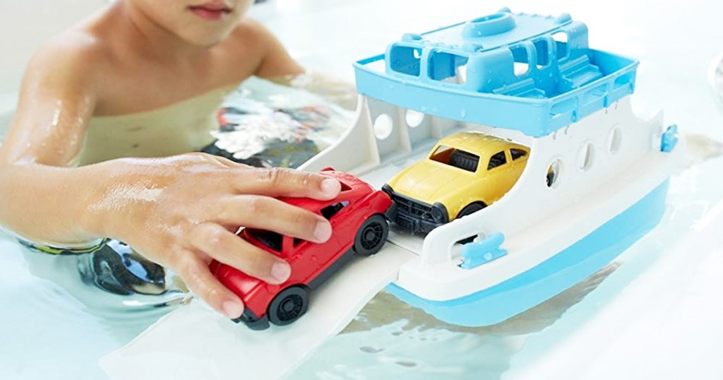 child in bathtub playing with blue and white ferry boat toy and holding red car in hand