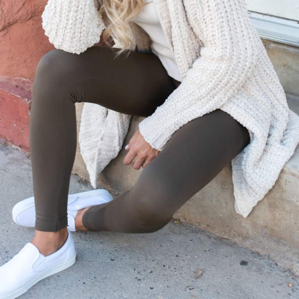 woman wearing coffee brown jane leggings sitting on cement bench wearing white shoes and cream cardigan 